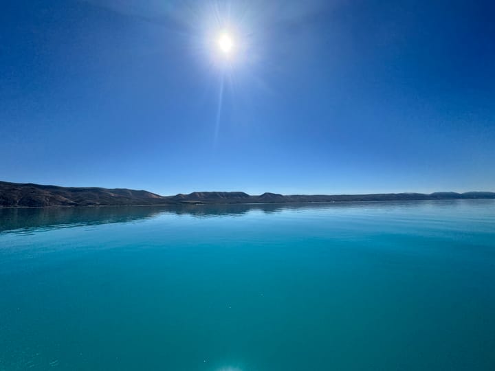 photo of crystal blue, clear water with no waves, with subtle mountains in the background and a sun's glare